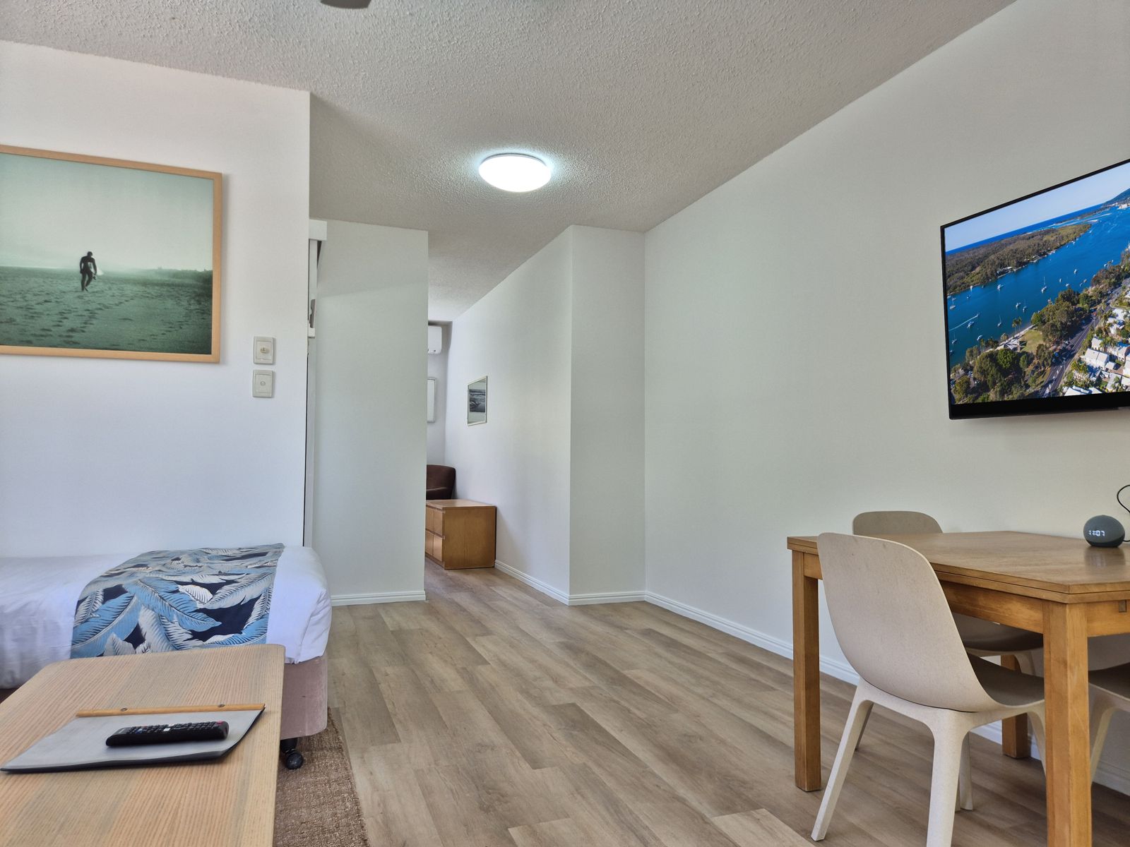 Open living area with single bed, dining table and wall-mounted Smart TV, looking through to the bedroom